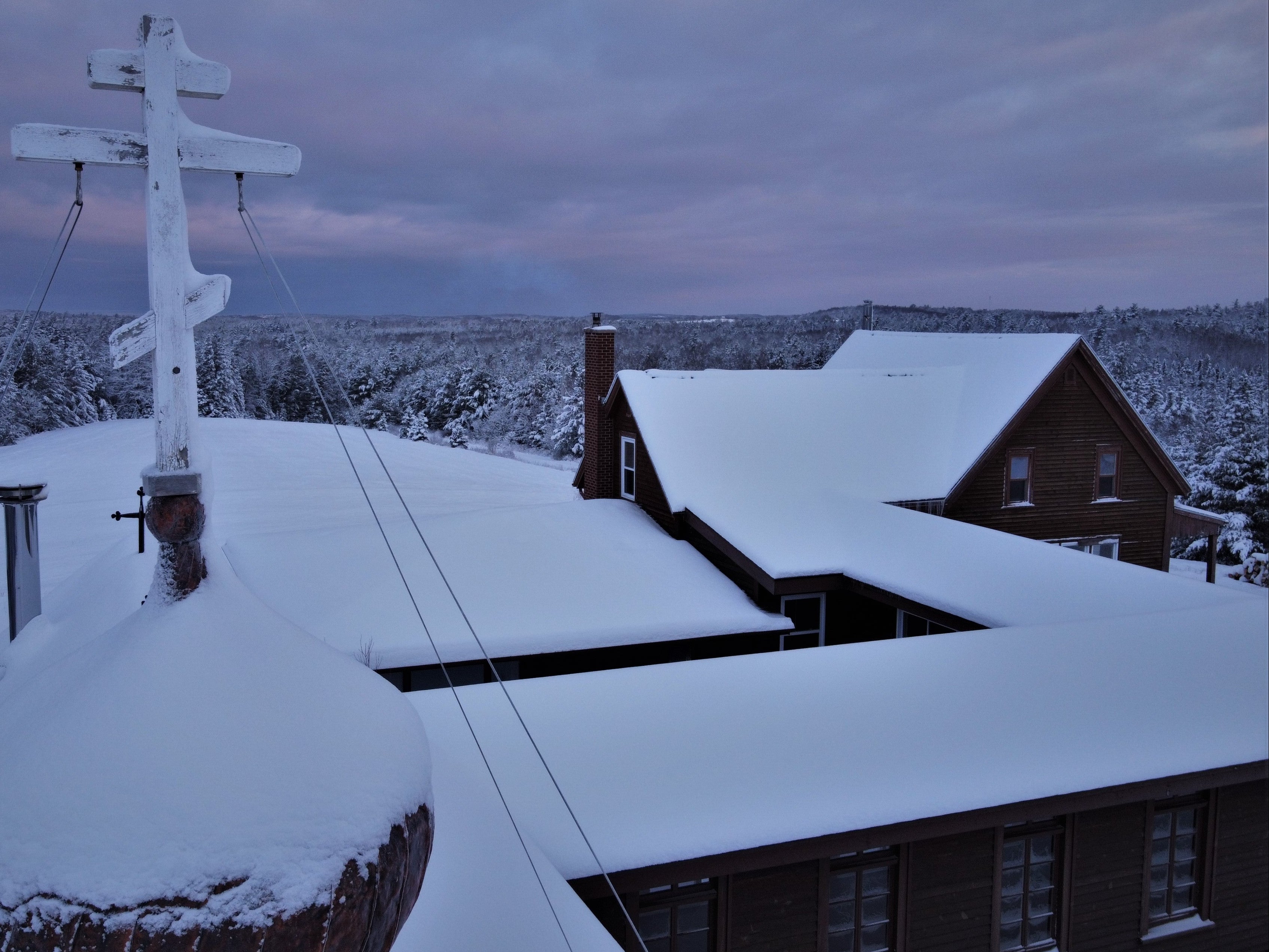 Snow-covered roof with a bell tower and a building in the background under a purple sky.