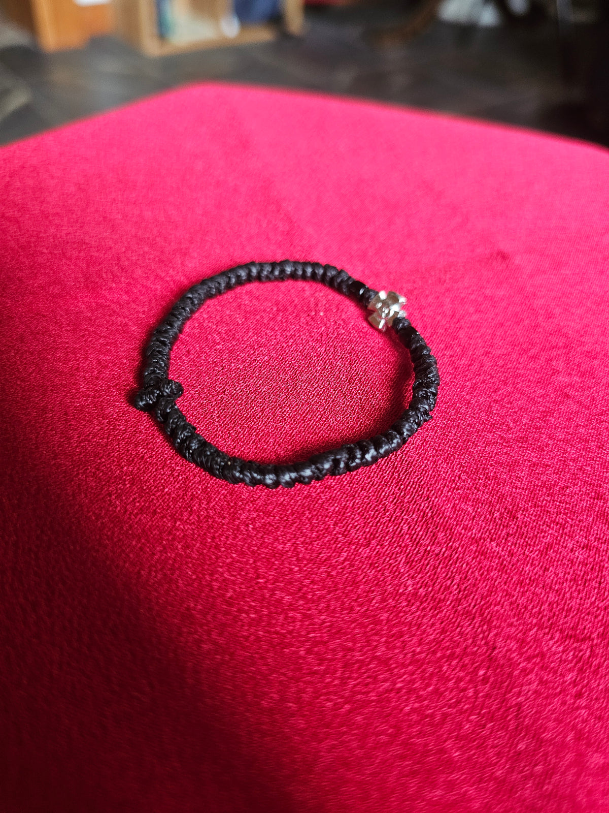 A black braided cord bracelet with a small silver bead accent, resting on a textured bright pink surface.